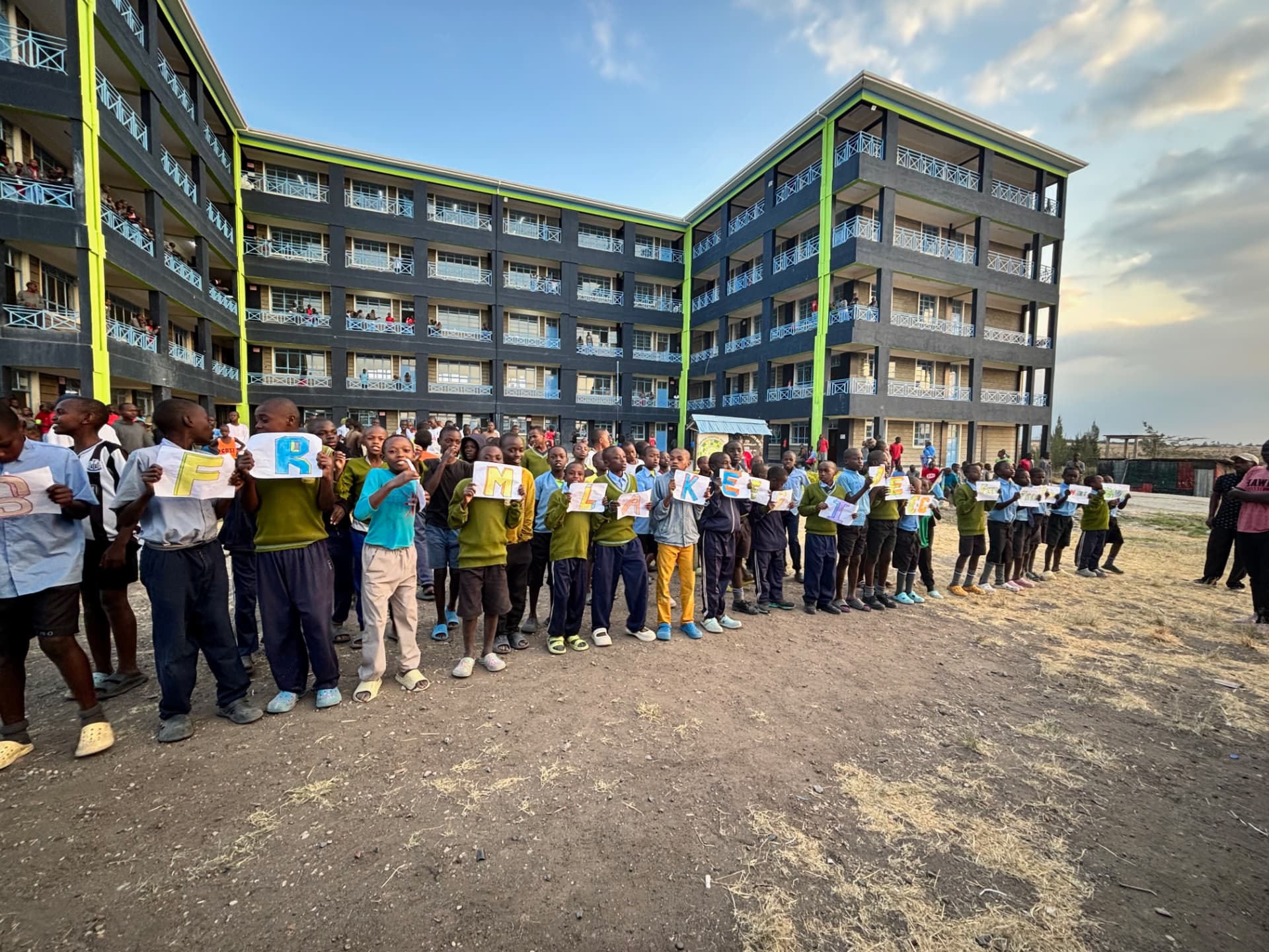 Students at Ndovoini School holding hand-lettered welcome signs in front of their classroom buildings